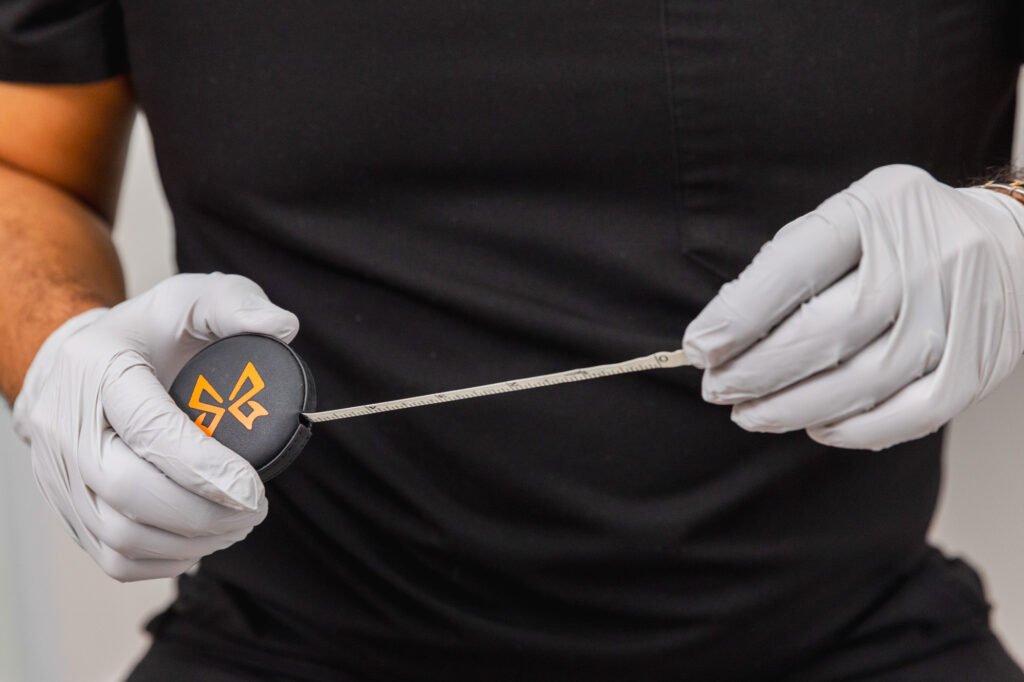 Close-up of a specialist in black scrubs and white gloves holding a measuring tape, preparing for a stomach fat removal consultation.