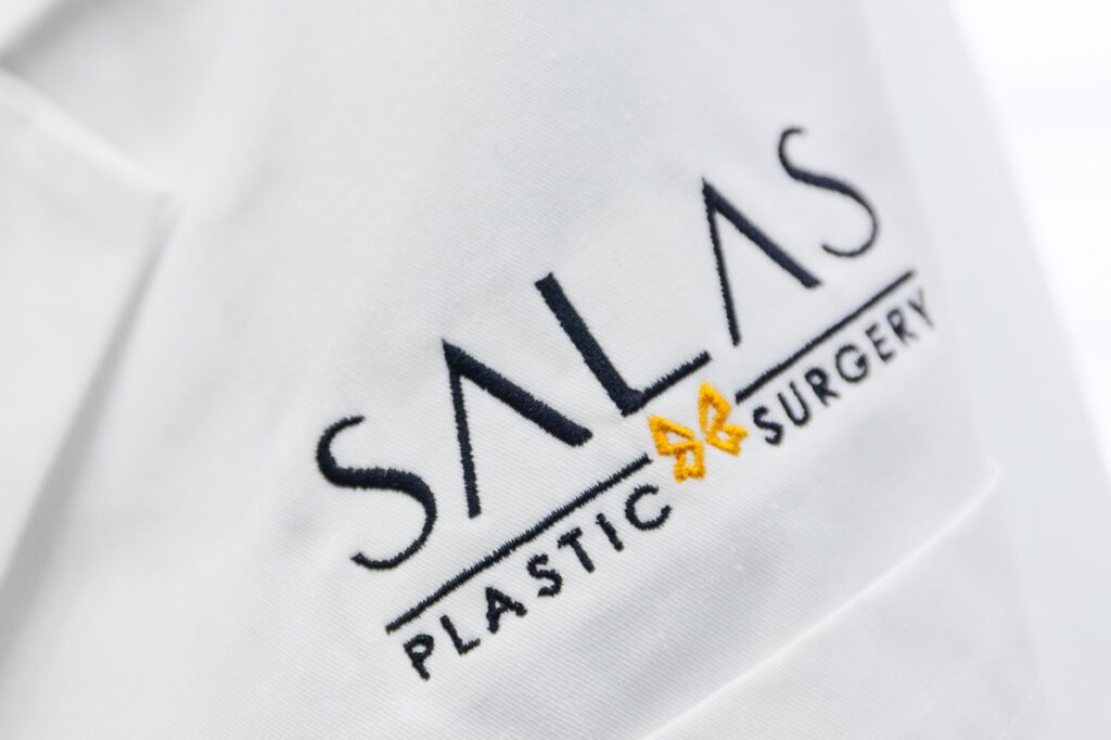 Close-up of the Salas Plastic Surgery logo on a white medical coat, representing the destination for expert care for chin lipo in Pembroke Pines.